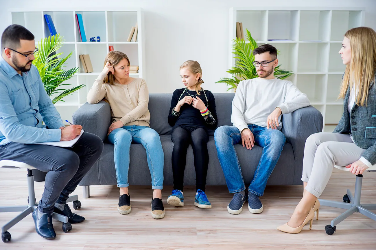 Five people sitting on a gray couch in a circle during a group therapy or support group session in a bright office with white shelving and potted plants.