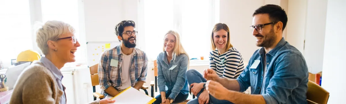 Five people sitting together in an office setting, smiling and engaged in conversation in a bright, modern workspace.