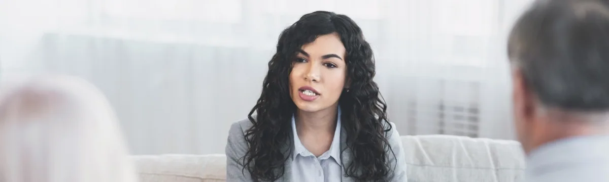 Woman with long black wavy hair wearing a light blue shirt seated in an office setting during a conversation