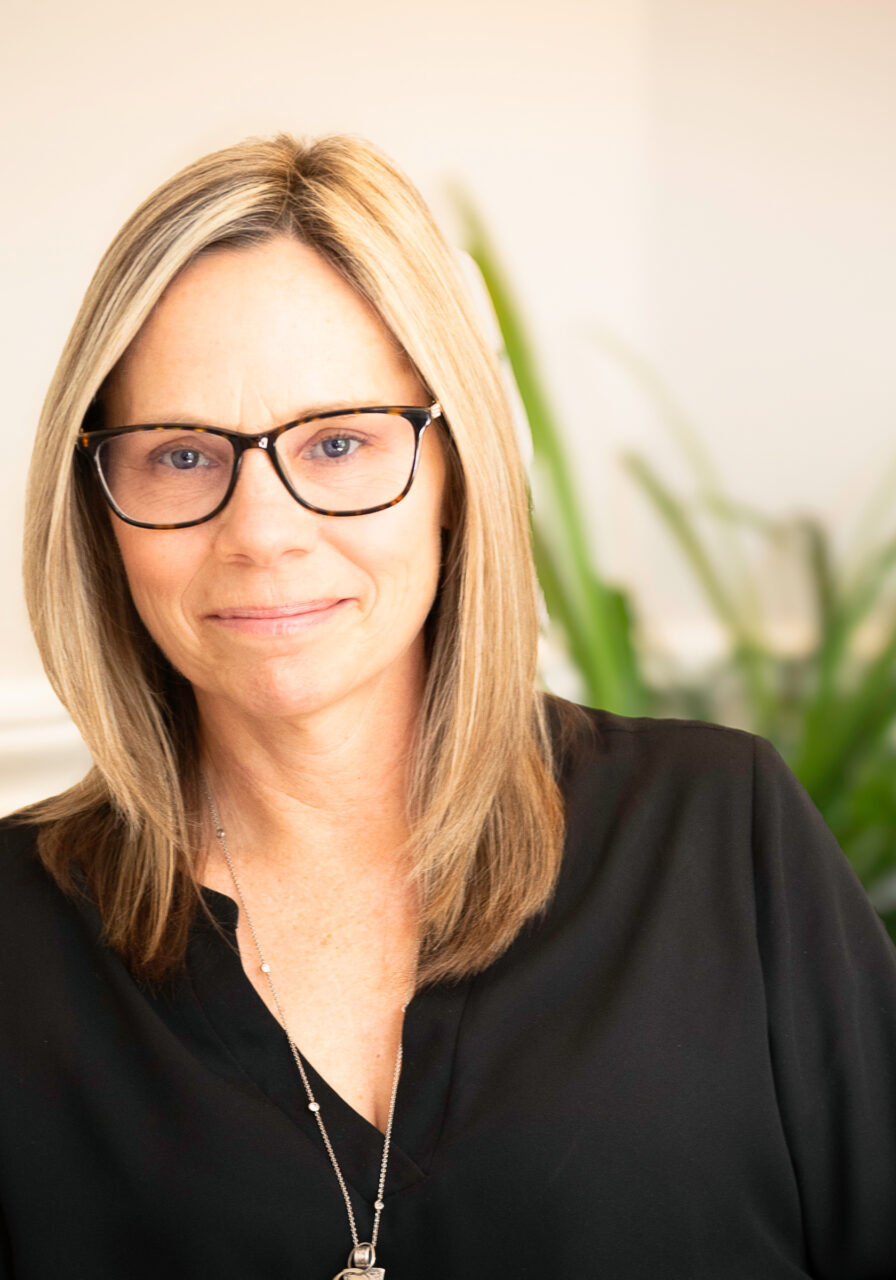 Woman wearing glasses and black shirt seated indoors with potted plant in background